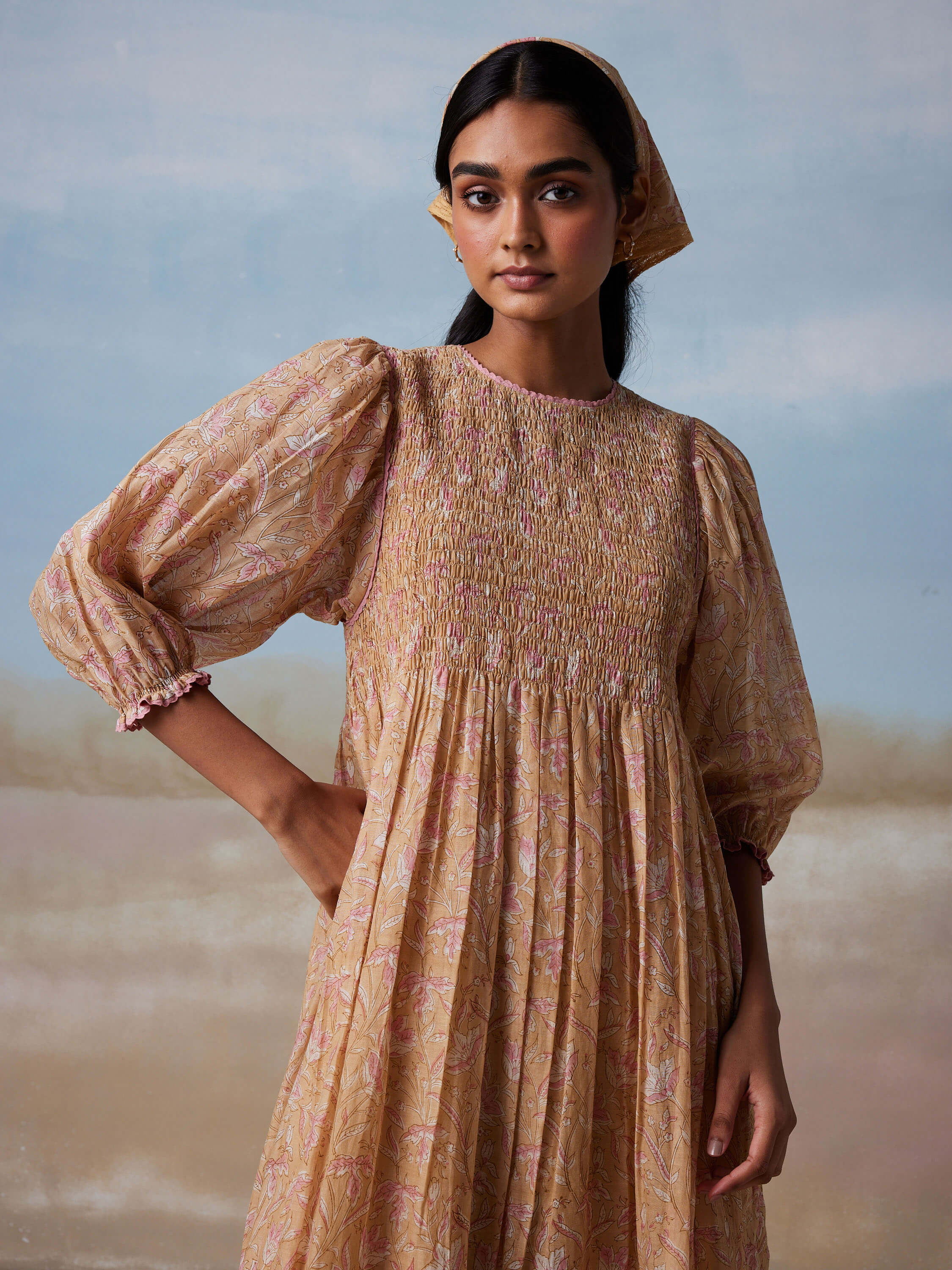 Woman in patterned long-sleeve dress with headscarf in front of pastel background.