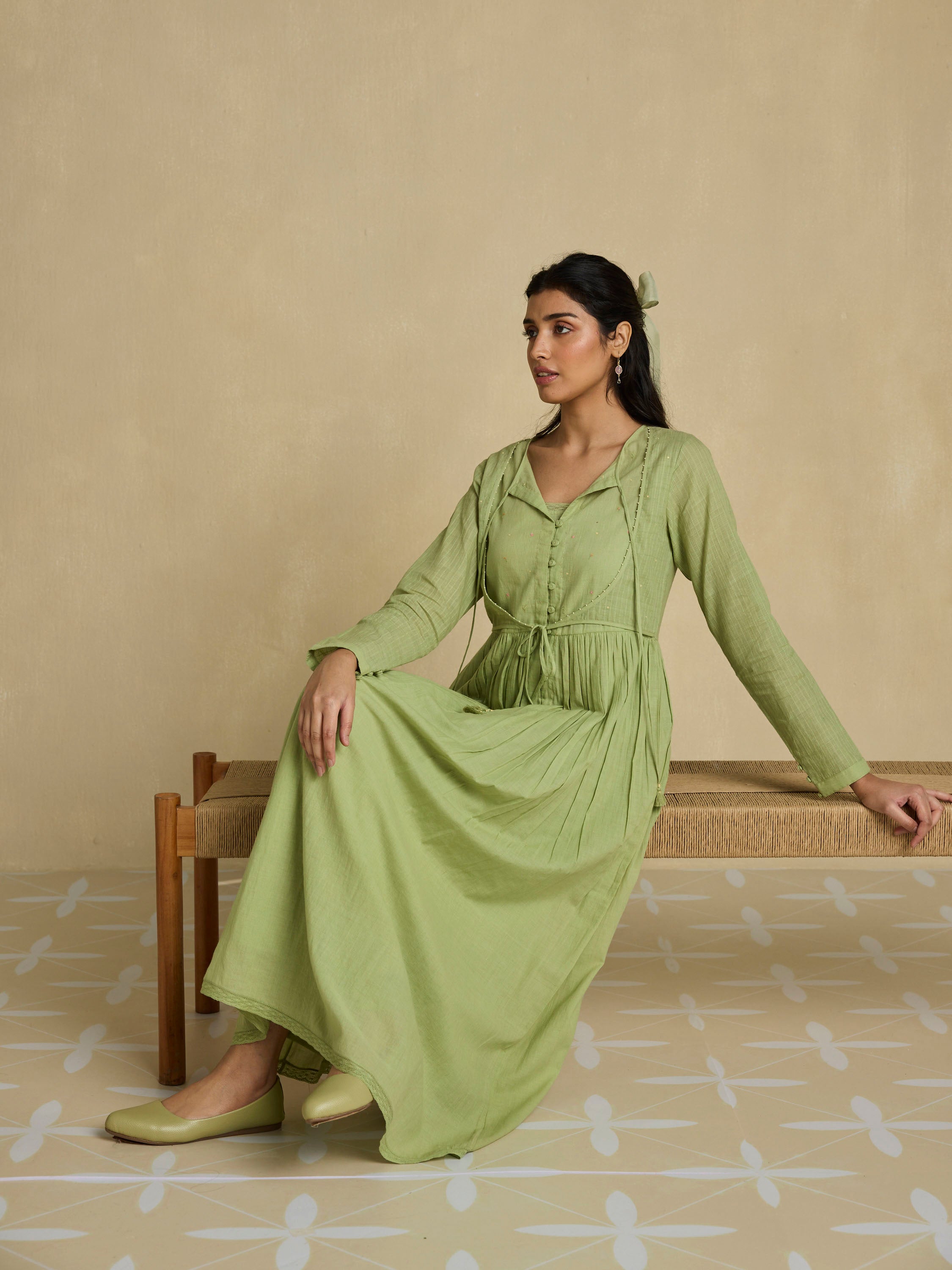 Woman in a green dress sitting on a wooden bench against a beige wall.
