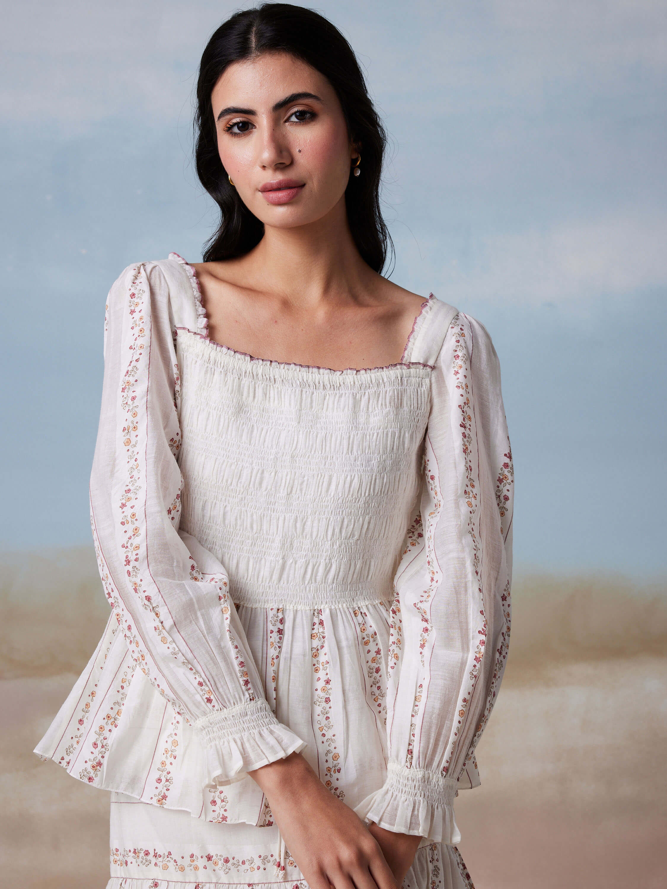 Woman in white floral-patterned blouse posing in front of a painted backdrop.