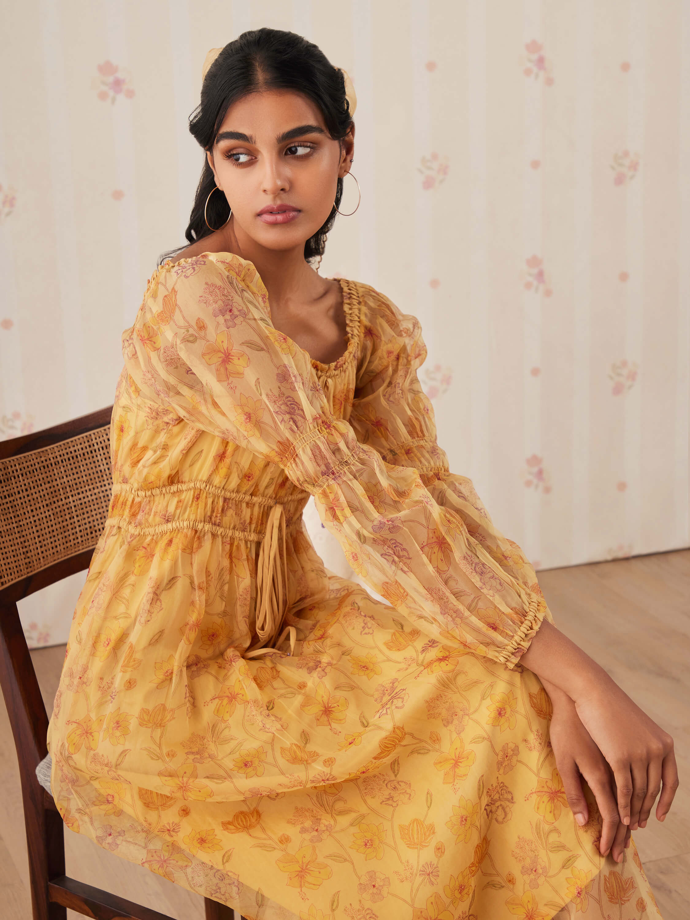 Woman wearing a yellow floral dress, sitting on wooden chair.