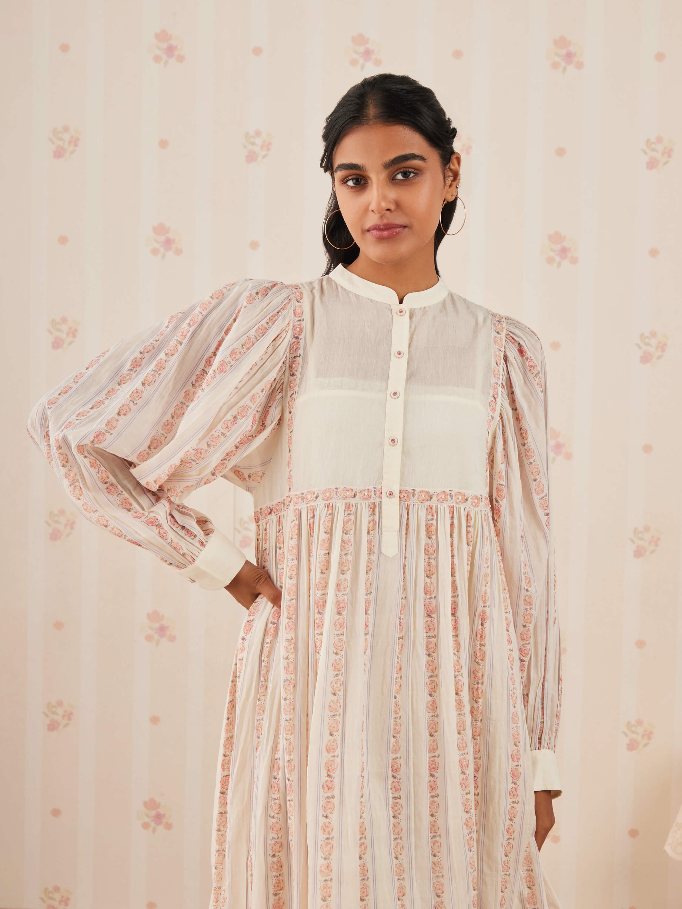 Woman in a floral patterned dress posing against a floral wallpaper background.