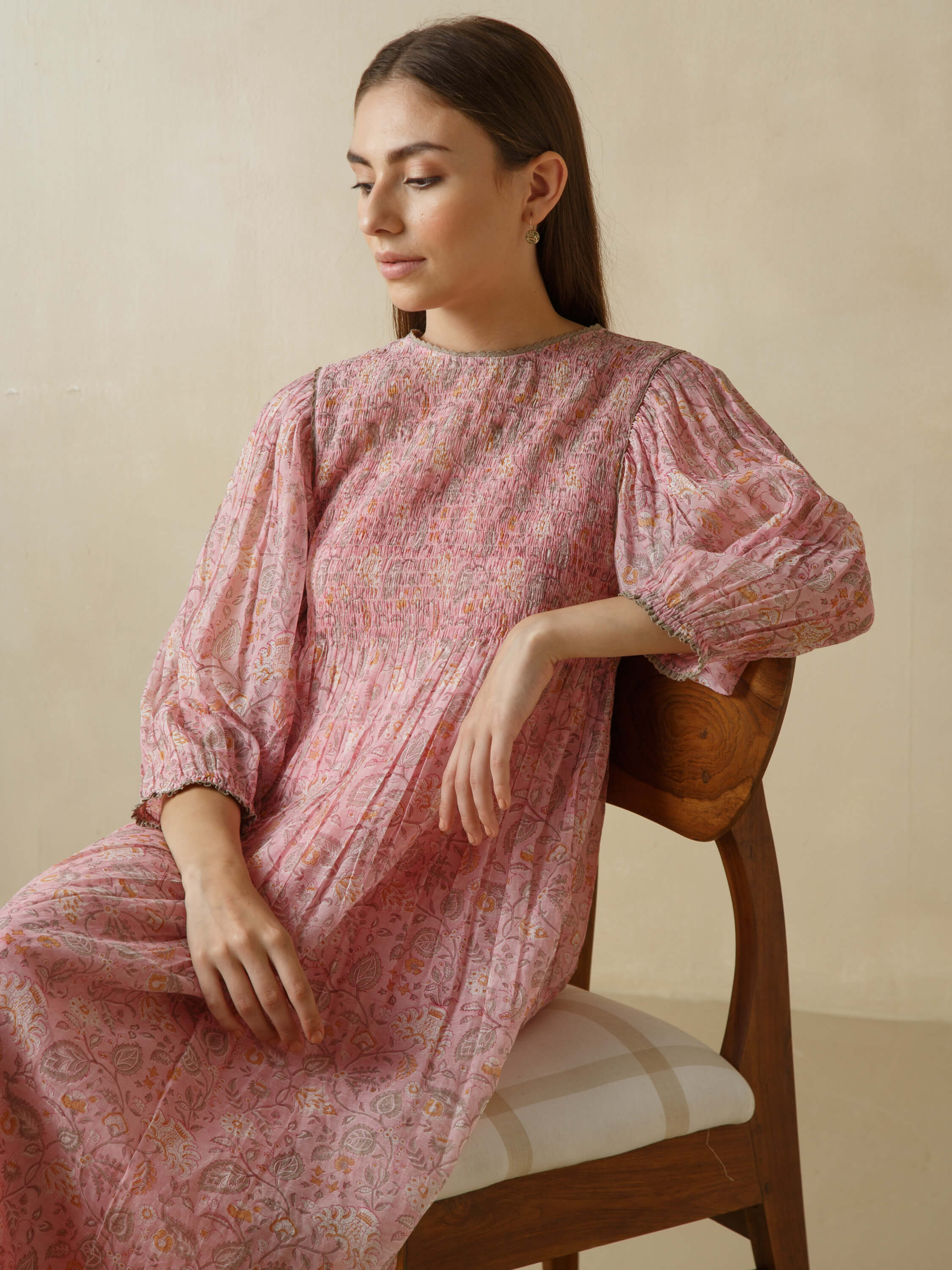 Woman in pink floral dress sitting on a wooden chair.