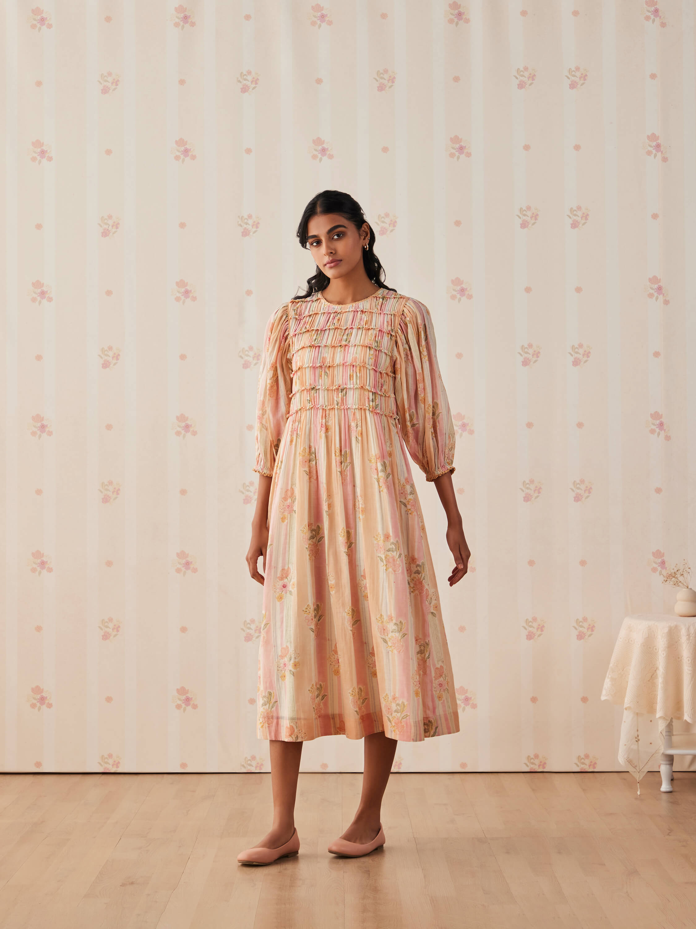 Woman in pink floral dress standing against patterned vintage wallpaper.
