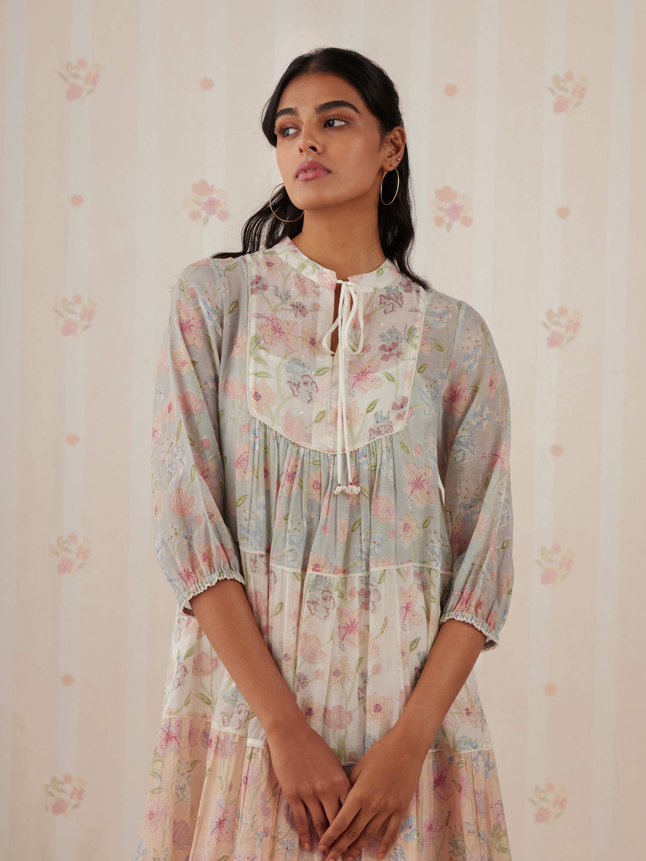 Woman wearing floral print dress, standing against striped floral wallpaper.