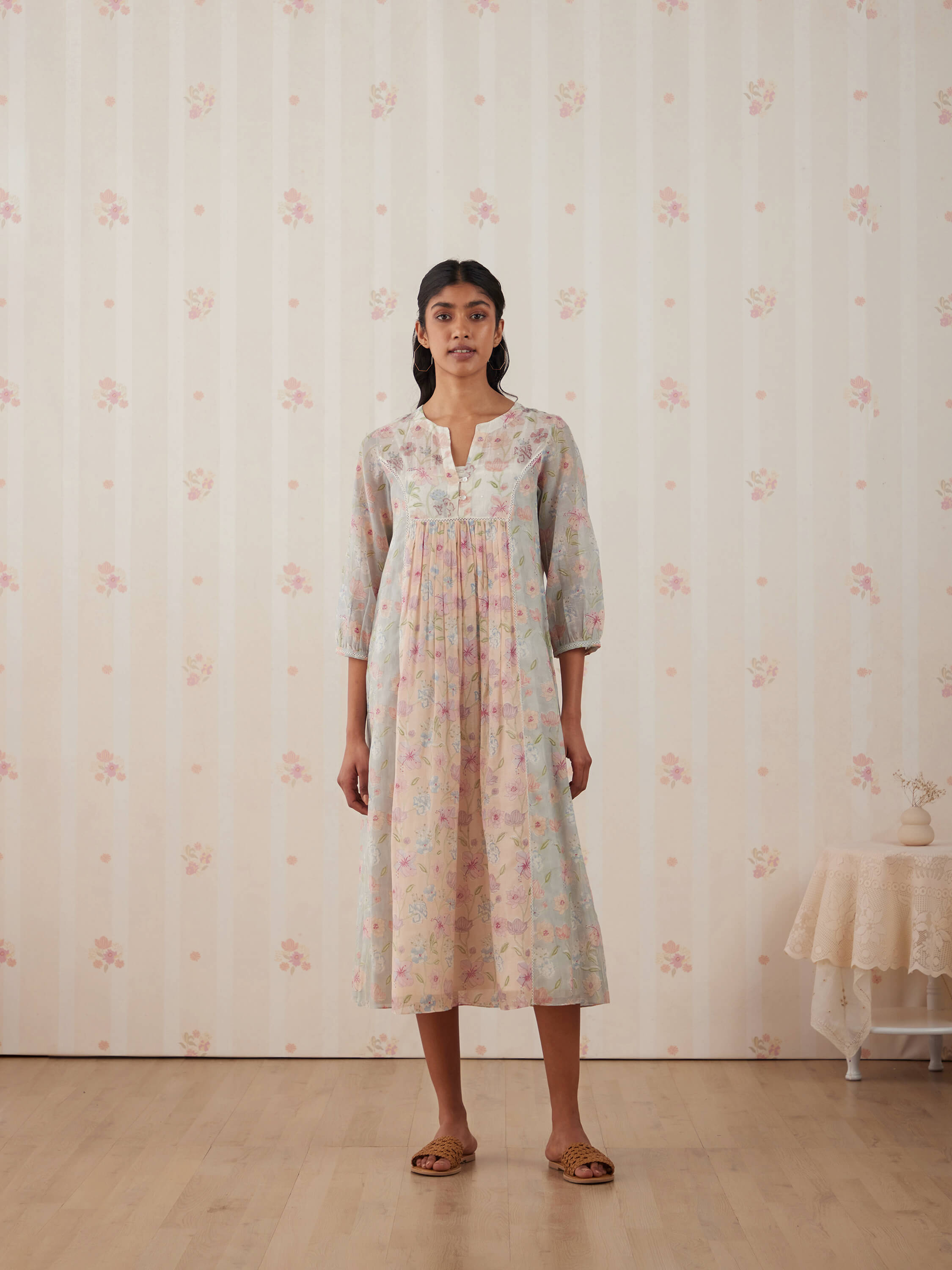 Woman in floral dress standing against floral wallpaper background.