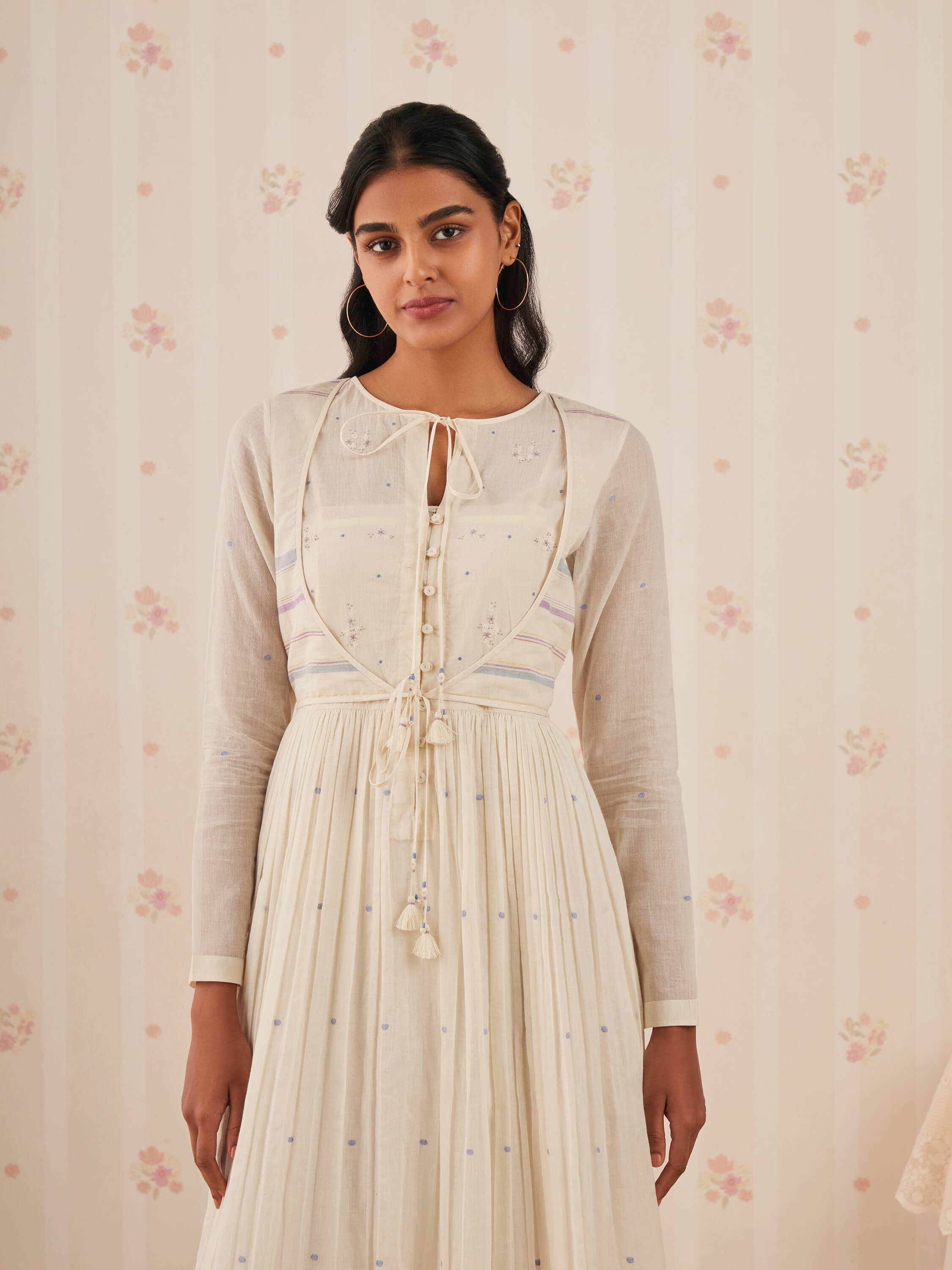 Woman wearing a cream ethnic dress with floral wallpaper background.