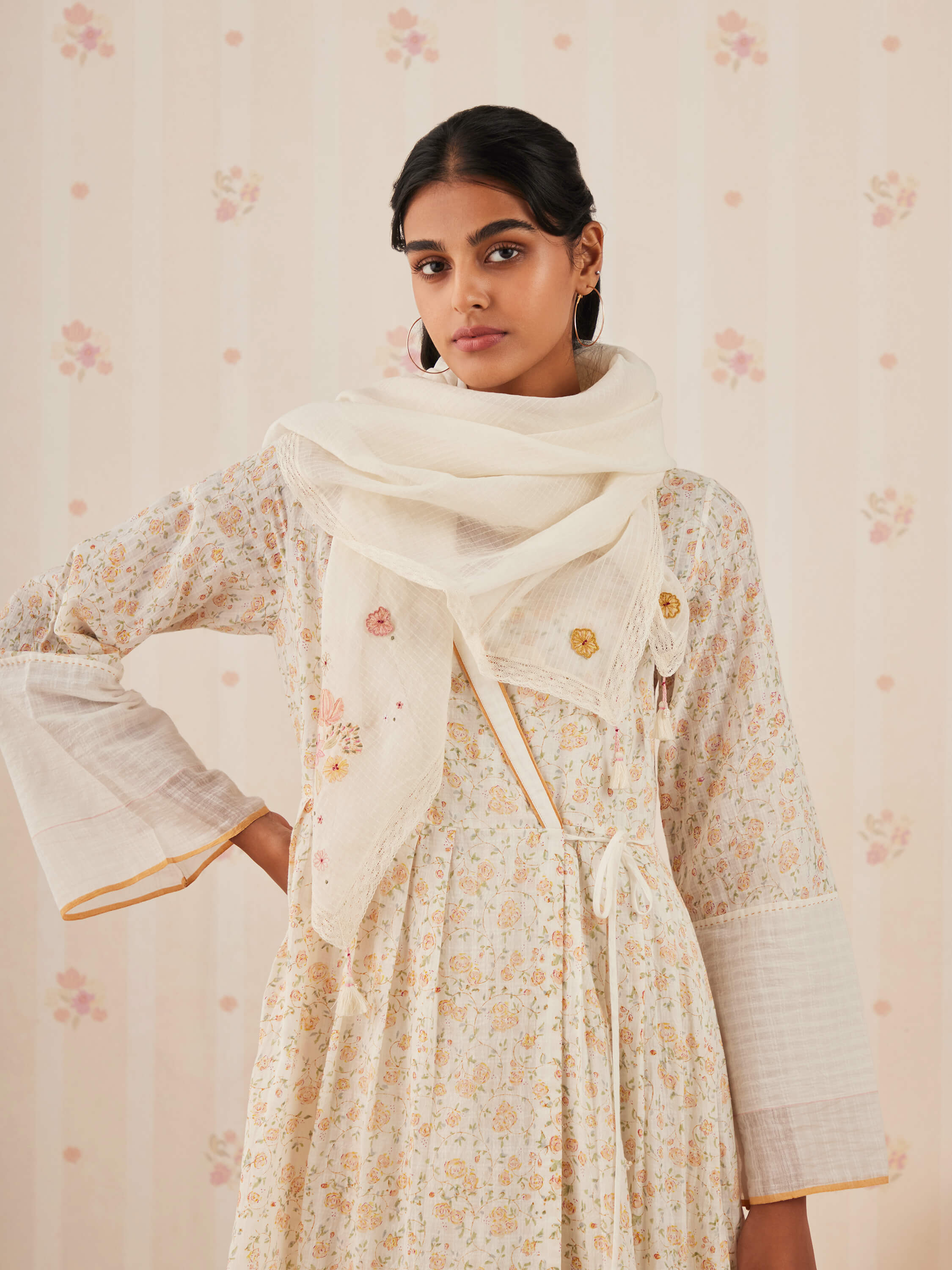 Woman in floral dress and white shawl against floral wallpaper.