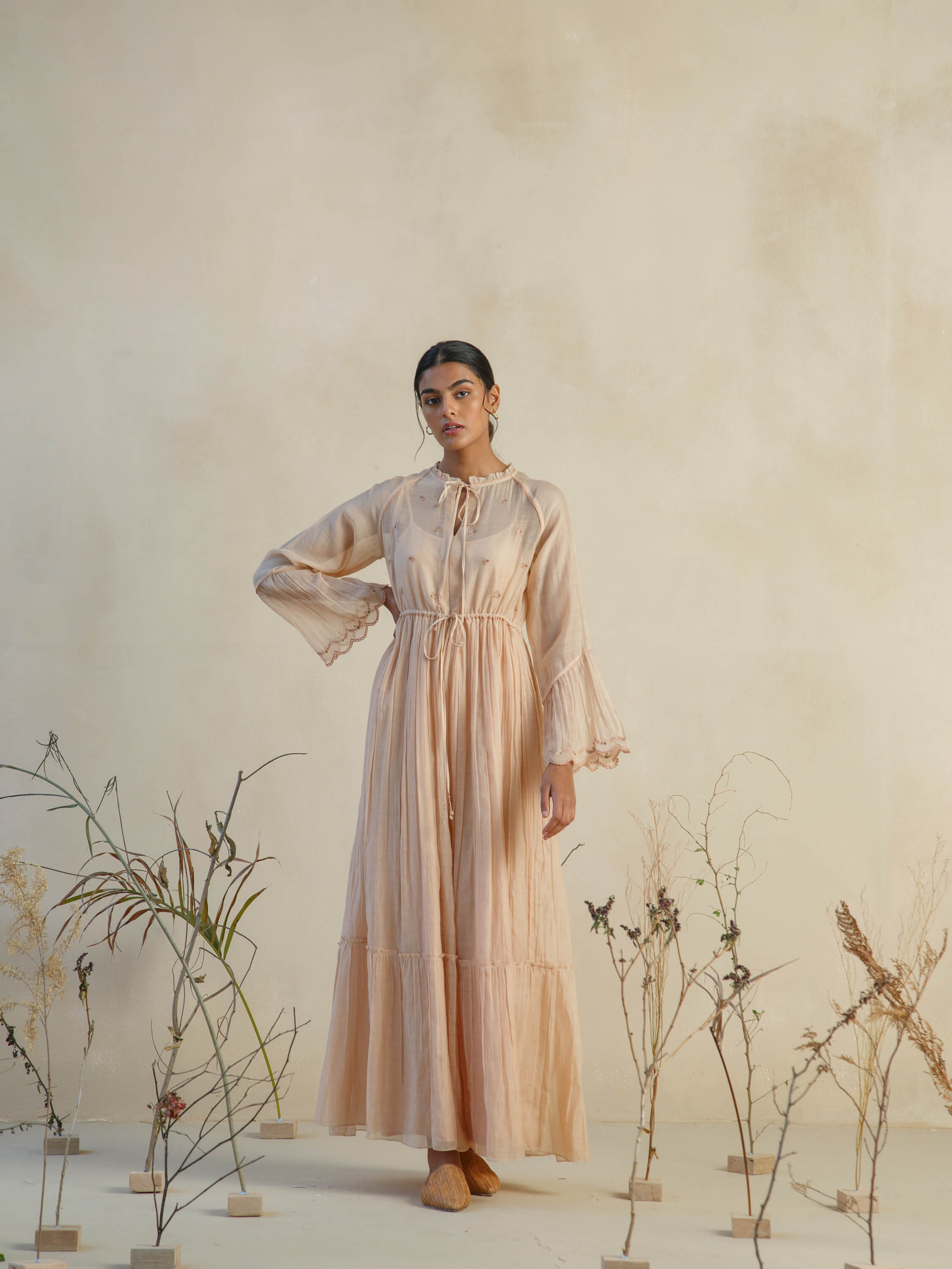 Woman in a light brown flowy dress surrounded by dried plants.