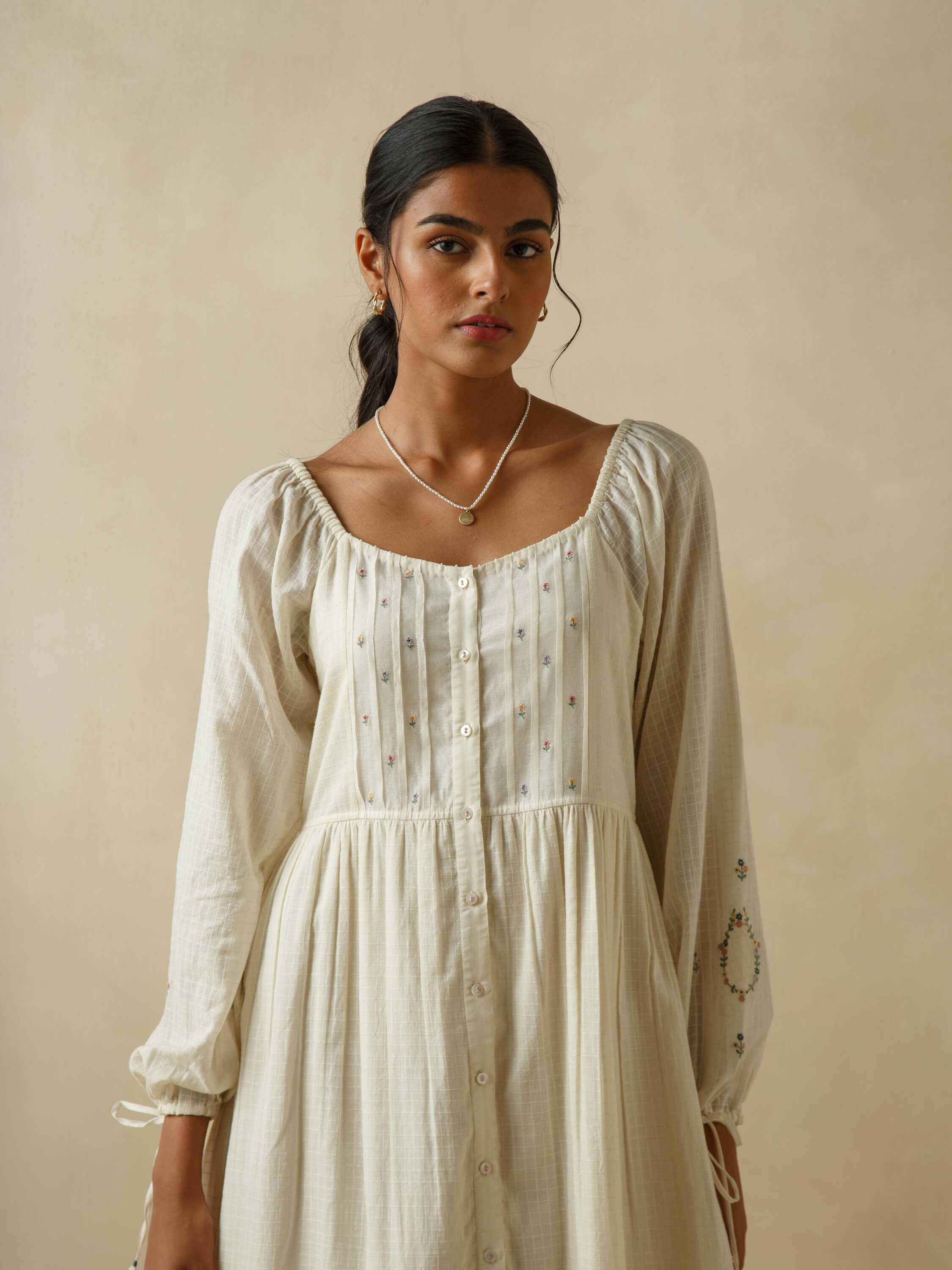 Woman in a beige dress with embroidery, wearing a necklace.