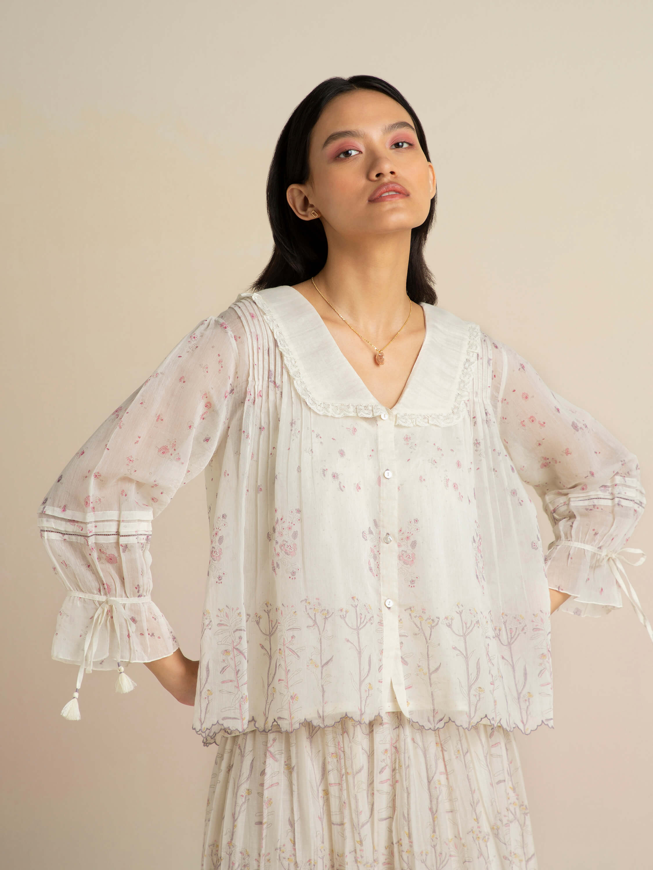 Woman in floral sheer blouse and skirt standing against beige background.
