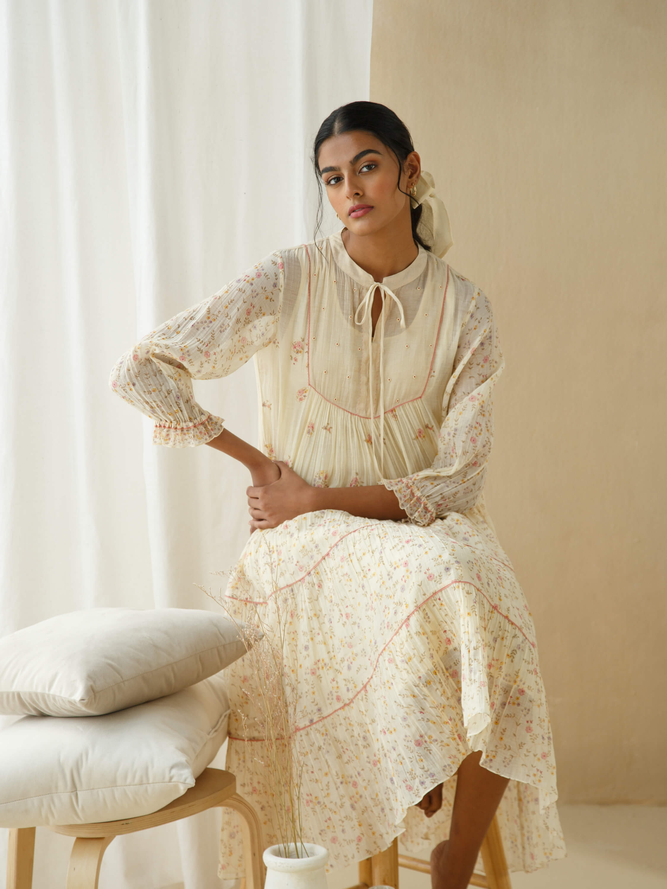 Woman in elegant floral dress sitting beside pillows with soft lighting.