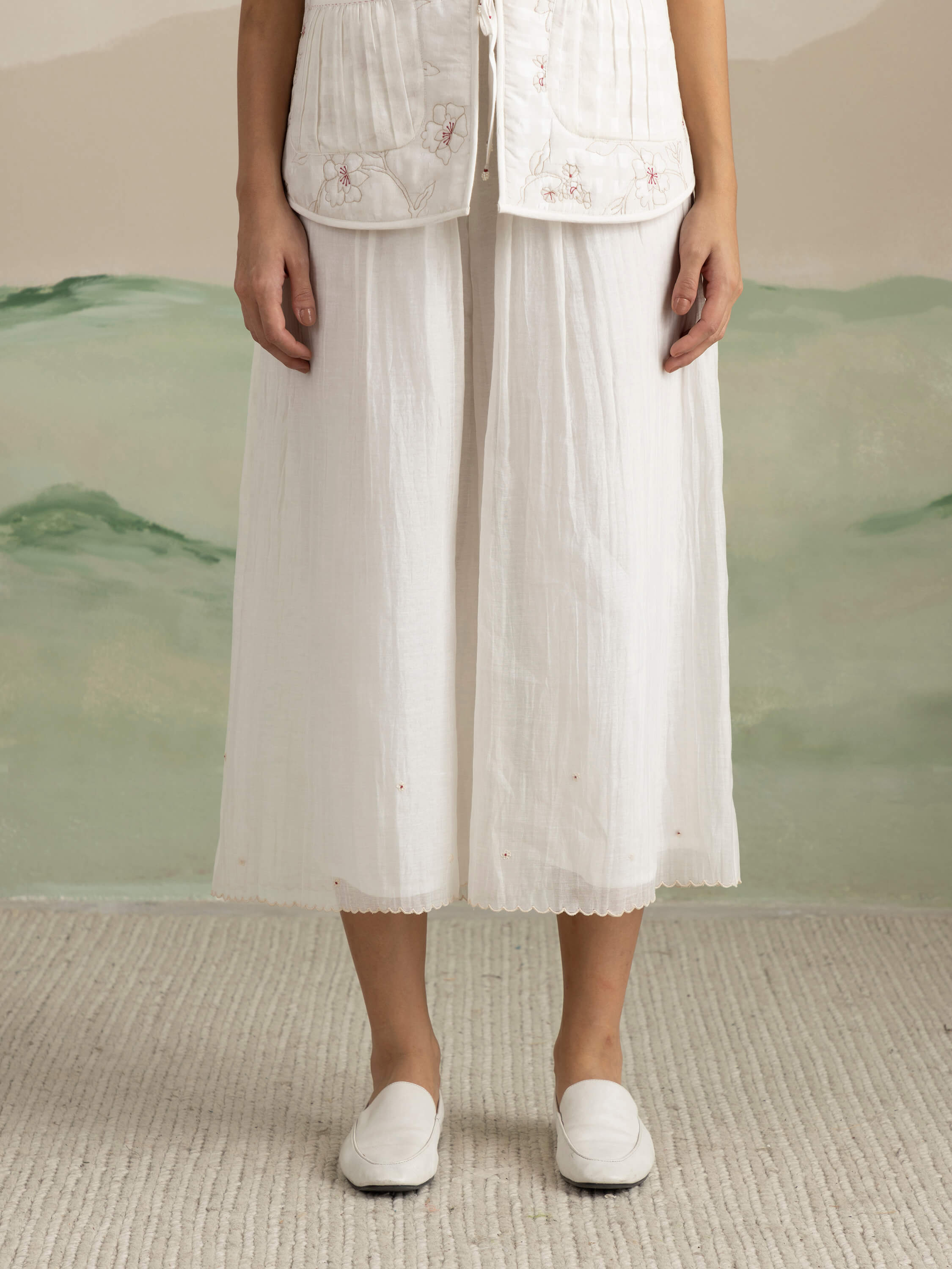 Woman in embroidered white pants and matching shoes against textured background.