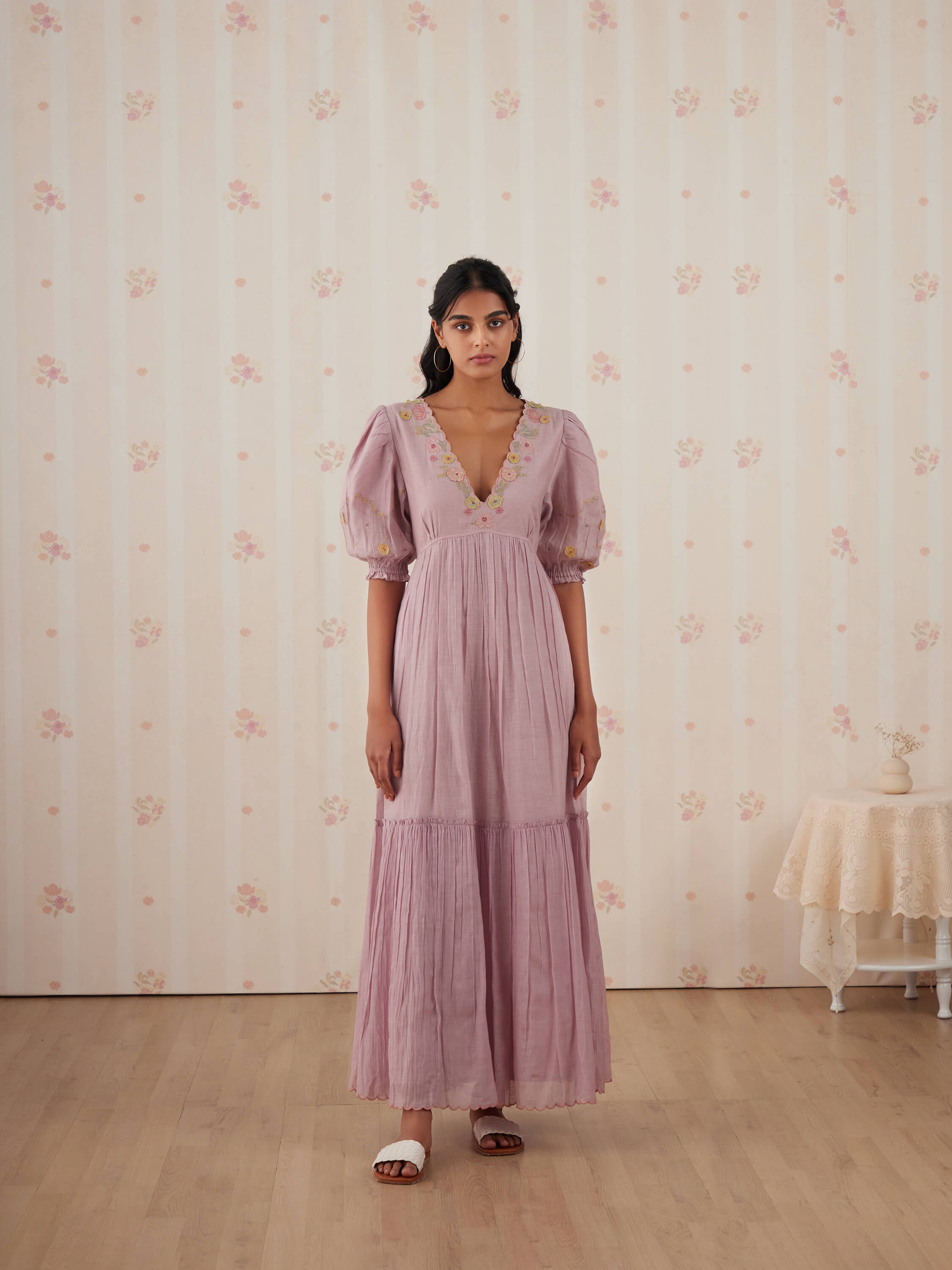 Young woman in pink dress stands against floral wallpaper background.