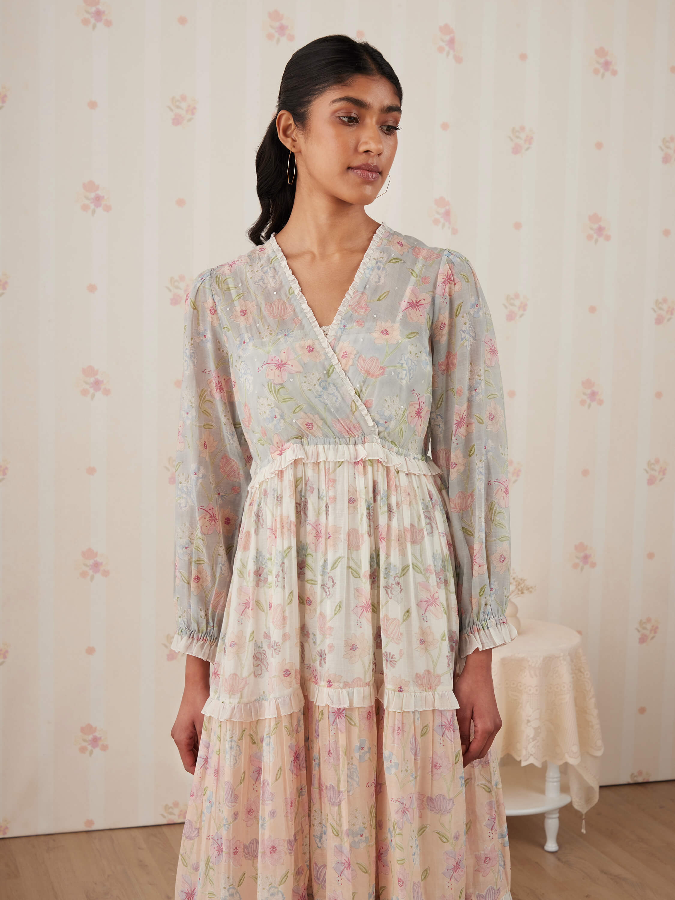 Woman in floral dress, standing against pastel flower wallpaper.